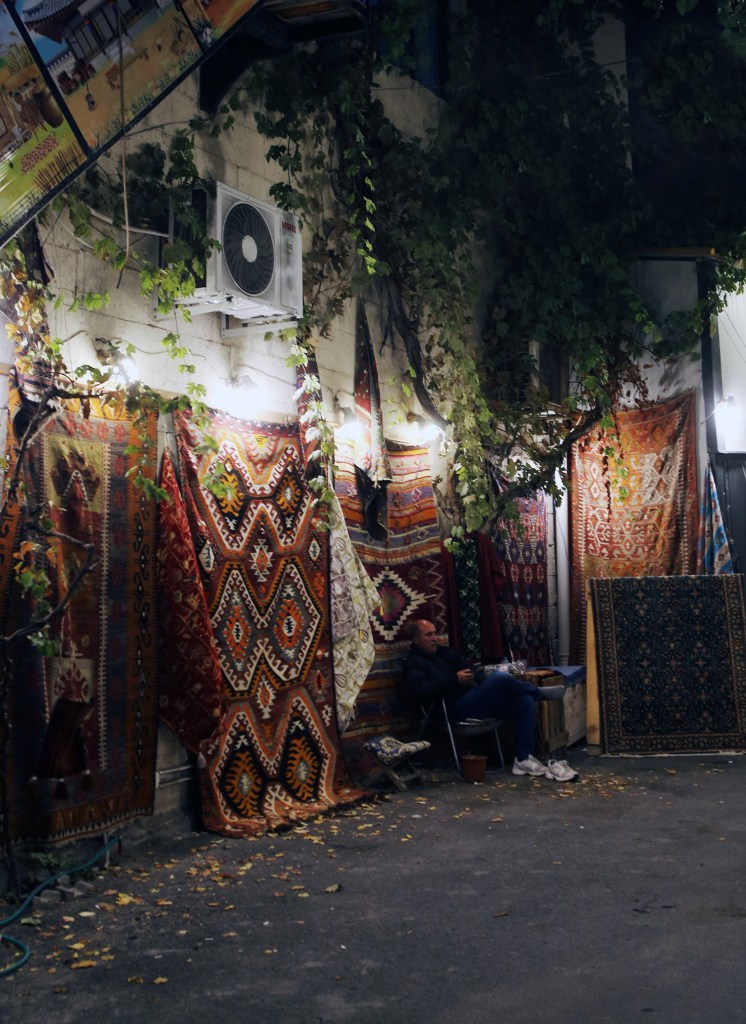 A street scene in Goreme, Cappadocia, featuring colorful carpets hanging on a wall, with a person sitting on a chair nearby.